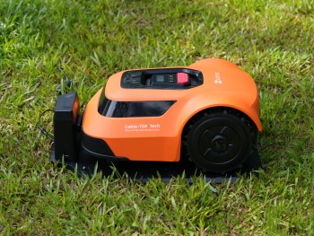 Ezviz RM600 robotgrasmaaier op het gras Oranje robotgrasmaaier op het gras, zwarte wielen en een laadstation ernaast.