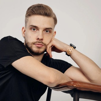 Havu Watches Apple Watch-bandje in berk elegant in gebruik. Een jonge man leunt tegen een lederen stoel terwijl hij een zwart t-shirt draagt en een houten Apple Watch-bandje in berkenkleur om heeft.