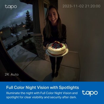 TP-LINK TAPO D210 deurbelcamera in nachtzichtmodus tijdens de schemering. Een vrouw staat in de deuropening tijdens de schemering, met een laagjestaart in haar hand. De tuin en verlichting van huizen zijn zichtbaar in de achtergrond.